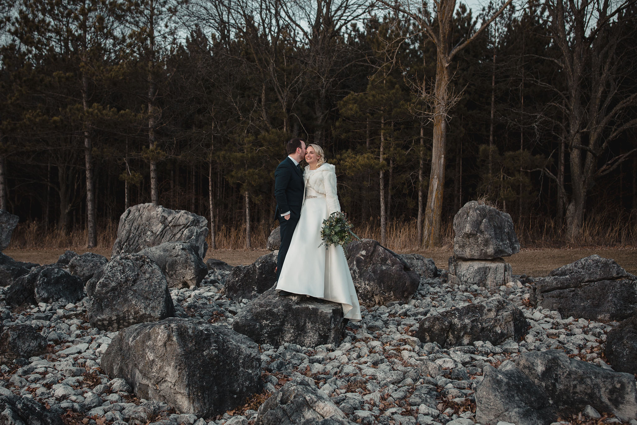 Winter wedding , couple standing on large rocks in front of lake. Bride wearing floor length winter white wedding dress with greenery floral and white fur stole to keep warm. Groom wearing classic dark blue suit with tie.